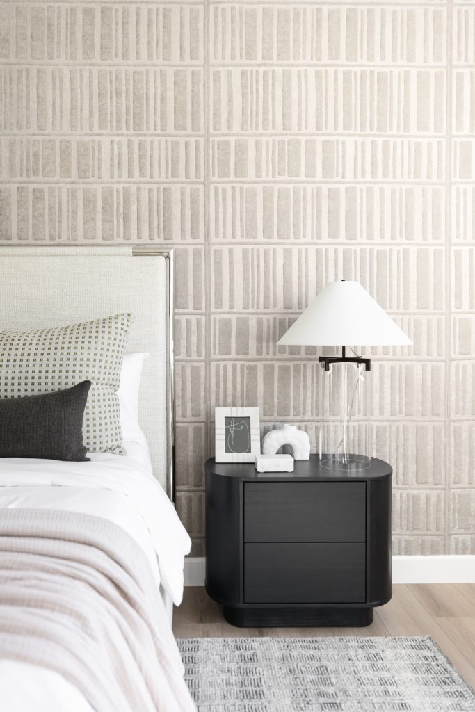 A bedroom features a beige patterned accent wall, a bed with neutral bedding, and a black nightstand with a lamp, framed art, and a decorative object. — Living with Lolo.