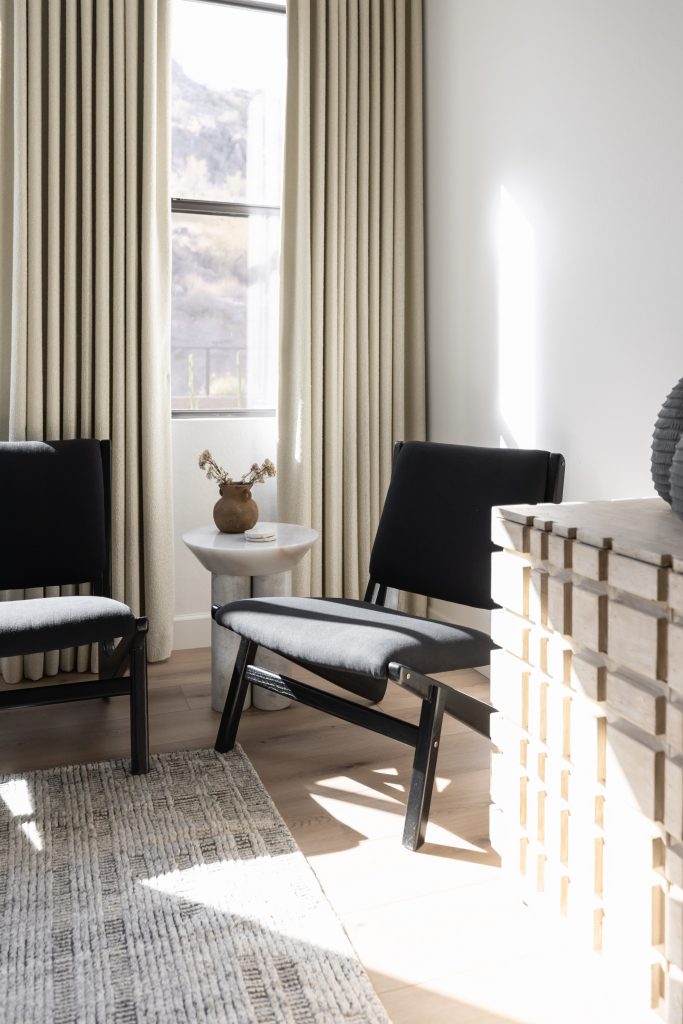 Two black chairs face a small side table holding a brown vase of dried flowers, positioned next to a window with beige curtains in a bright, modern room. — Living with Lolo.