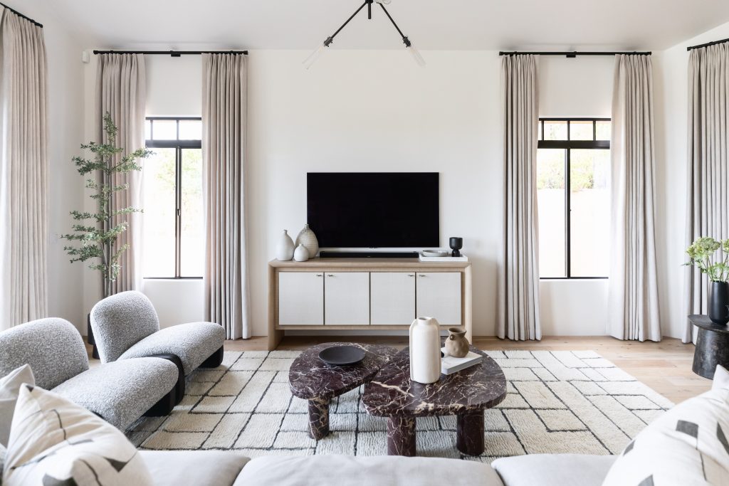 Modern living room featuring neutral tones, a wall-mounted TV above a console, two marble coffee tables, a patterned rug, light gray sofas, and large windows with beige curtains—Living with Lolo.