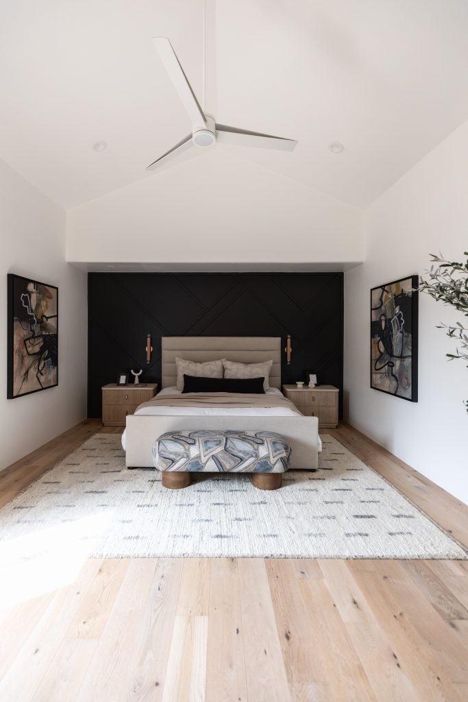 Modern bedroom featuring a neutral bed, abstract art on white walls, a black accent wall, wooden nightstands, a patterned bench at the foot of the bed, and light wood flooring—Living with Lolo.