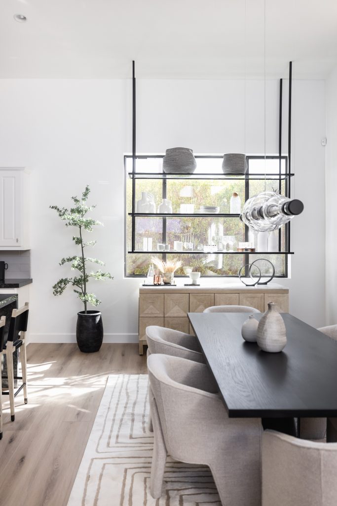 Modern dining room featuring a black table, light gray chairs, minimalist decor, a large window with shelves, pendant lights, and a potted plant in the corner—Living with Lolo.