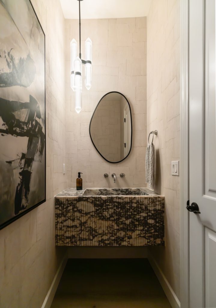 Narrow bathroom featuring abstract wall art, a marble-patterned sink, an oval mirror, a towel ring, a soap dispenser, and modern hanging lights—Living with Lolo.