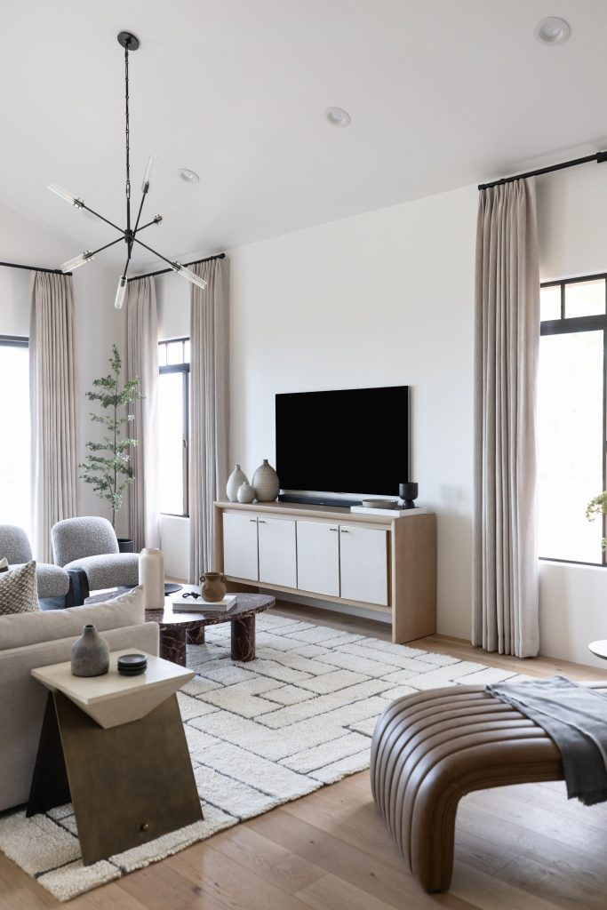 Modern living room with neutral colors, a geometric chandelier, TV on a cabinet, gray chairs, patterned rug, large windows, and minimalist decor—Living with Lolo.