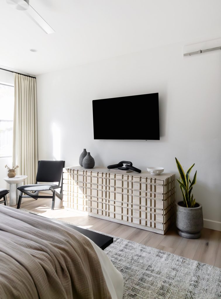 A modern bedroom displays a wall-mounted TV above a patterned dresser, two vases, a phone, a potted plant, and a black accent chair. Natural light enters through a window with sheer curtains—Living with Lolo.
