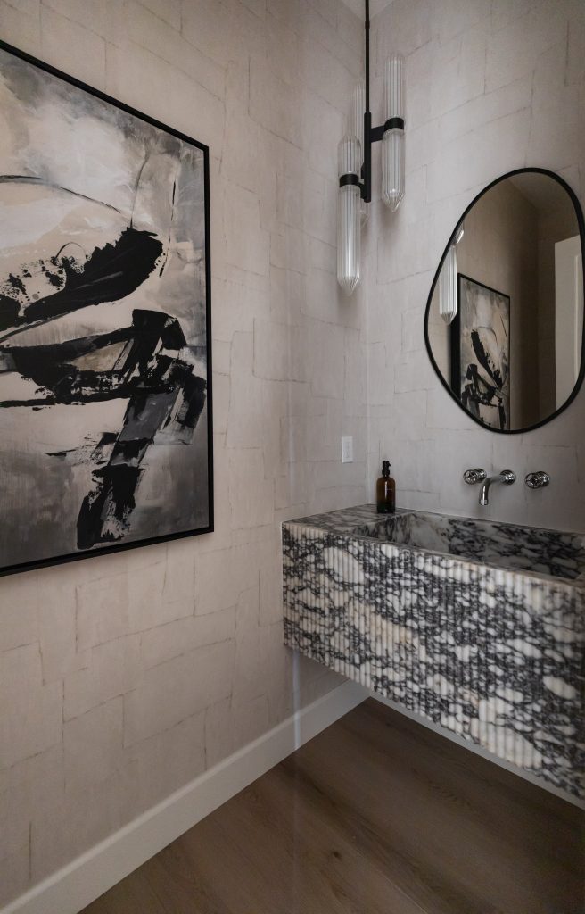 A modern bathroom with an abstract art piece, a marble-patterned floating sink, an oval mirror, a wall-mounted faucet, and two vertical pendant lights. — Living with Lolo.