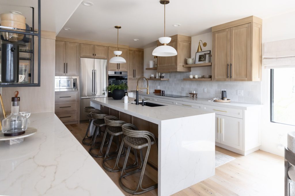 Modern kitchen featuring light wood cabinets, white marble countertops, a central island with four woven bar stools, stainless steel appliances, and pendant lights—Living with Lolo.