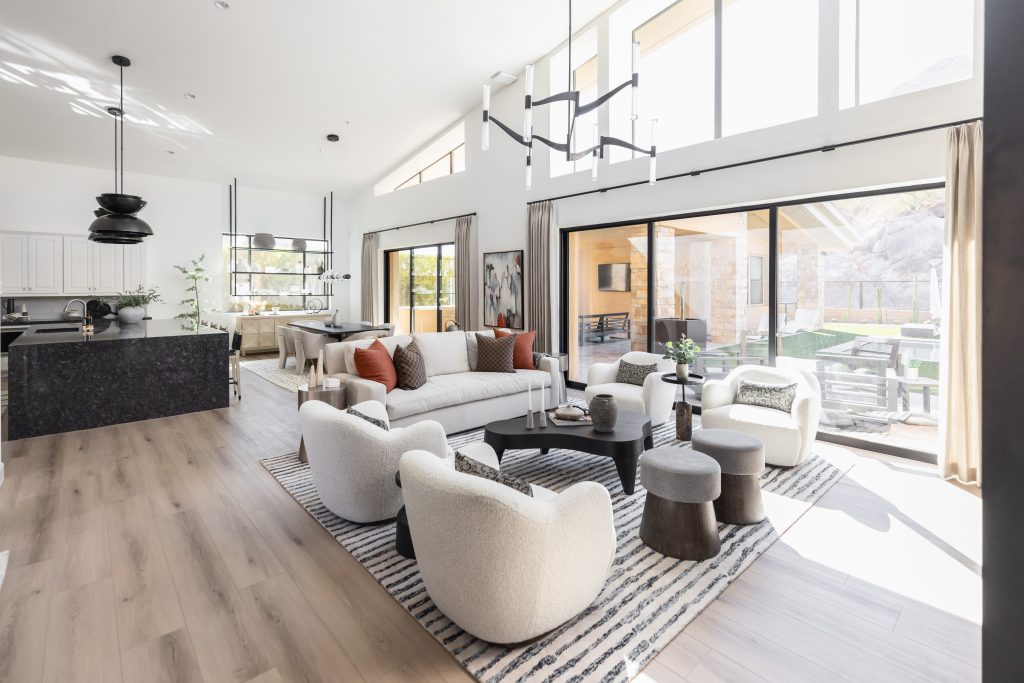 Modern living room featuring neutral furniture, a striped rug, large windows, and sliding doors to an outdoor patio. A kitchen with a dark island is visible in the background — Living with Lolo.