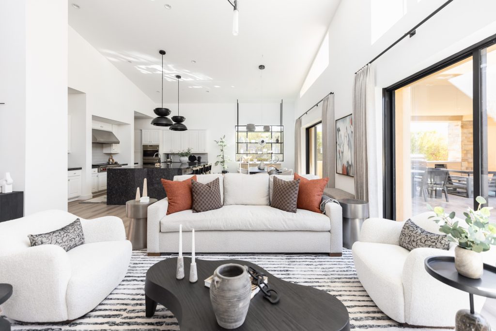 Modern living room featuring a beige sofa, two armchairs, and a black coffee table. Open kitchen and dining area are visible in the background with large windows allowing natural light.— Living with Lolo.