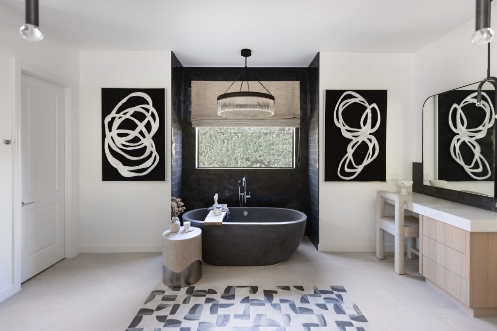 Modern bathroom with a black freestanding tub, abstract art on dark tiled walls, a round side table, and a light vanity on the right. A window is centered above the tub—Living with Lolo.