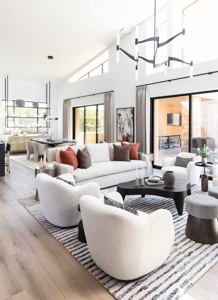 Modern living room with white sofas and chairs, a black coffee table, large windows, light wood floors, striped rug, and contemporary chandeliers. Dining area is visible in the background—Living with Lolo.