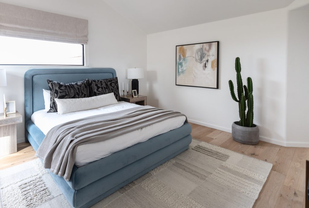 Modern bedroom featuring a blue upholstered bed with neutral bedding, two nightstands, a large potted cactus, abstract wall art, and a window with a gray shade—Living with Lolo.
