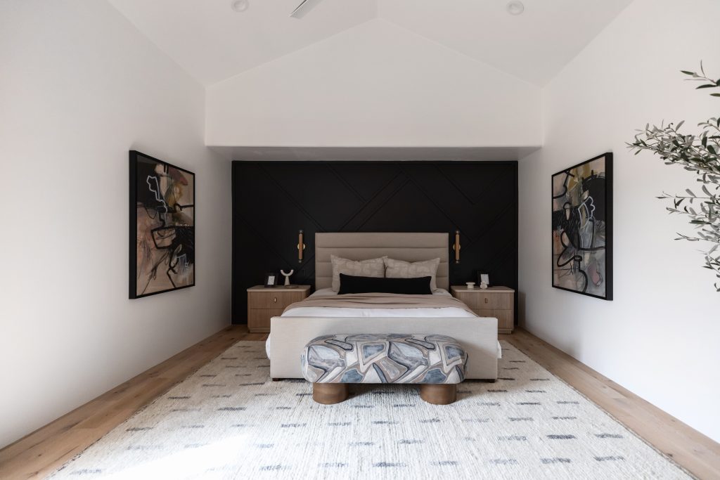 Modern bedroom featuring a beige bed, black accent wall, two abstract paintings, light wood nightstands, a patterned bench at the foot of the bed, and a neutral rug on wooden flooring.— Living with Lolo.