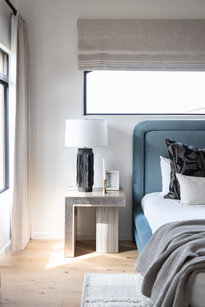 Modern bedroom featuring a blue upholstered bed with patterned pillows, a gray nightstand, black lamp, framed photo, and beige curtains by a large window—Living with Lolo.
