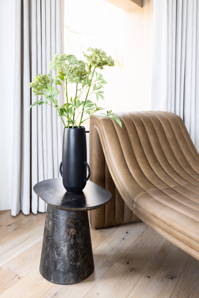 A tan leather chaise lounge sits next to a round black side table and a black vase with green flowers, all on light wood flooring in front of white curtains. — Living with Lolo.