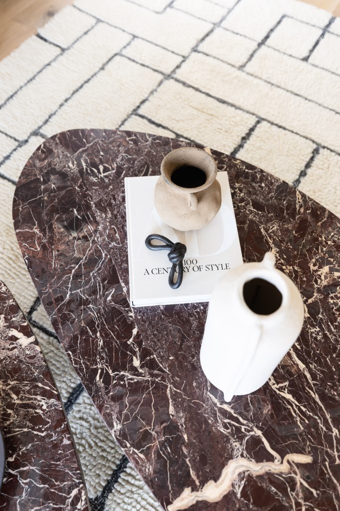 A brown marble coffee table sits on a patterned rug, holding a book, a small beige vase, a black decorative knot, and a white vase—Living with Lolo.