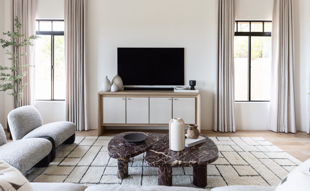 A modern living room features a TV on a console, two vases, a dark marble coffee table, light chairs, a patterned rug, and tall windows with sheer curtains. — Living with Lolo.