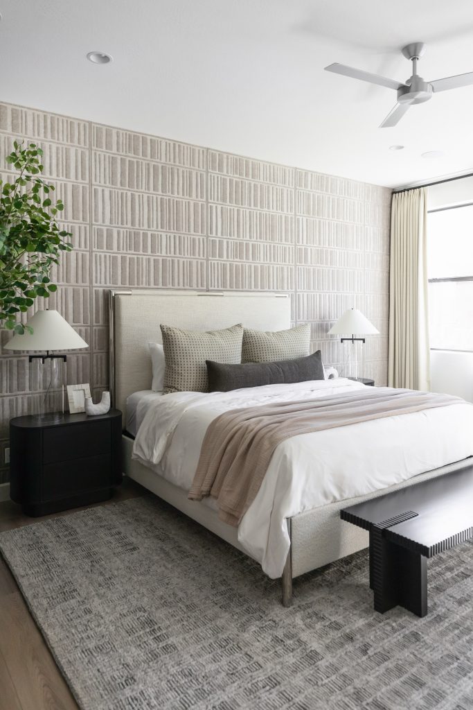 Modern bedroom with neutral colors, a double bed, patterned wallpaper, bedside tables with lamps, a black bench at the foot of the bed, a large gray rug, and a leafy plant in the corner near the headboard—Living with Lolo.