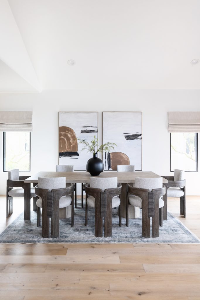 Modern dining room featuring a wooden table, eight cushioned chairs, a black vase with greenery, two abstract paintings on the wall, and neutral-toned decor—Living with Lolo.