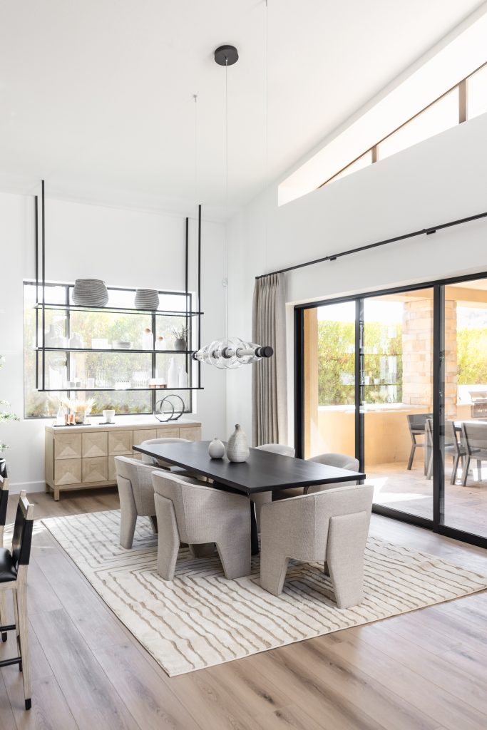 Modern dining room with a dark rectangular table, six light gray chairs, a striped rug, large windows, and contemporary pendant lighting above the table. — Living with Lolo.