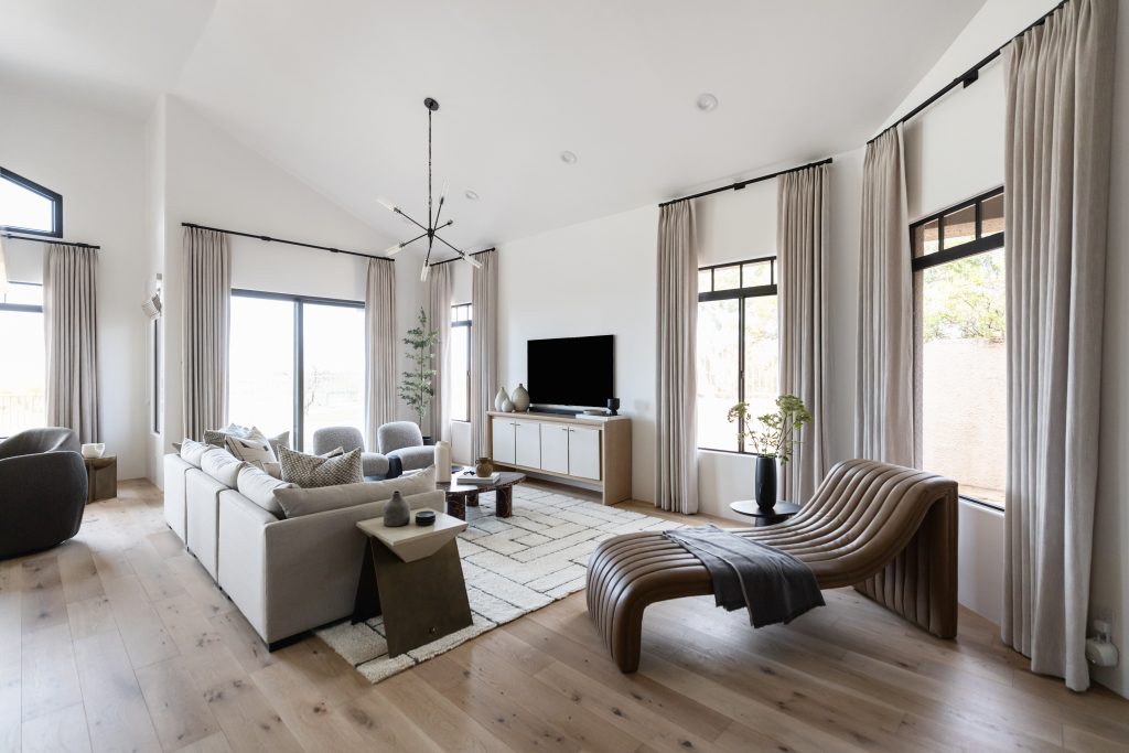 Modern living room featuring beige sofas, a wooden lounge chair, a TV on a cabinet, large windows with curtains, and light wood flooring — Living with Lolo.