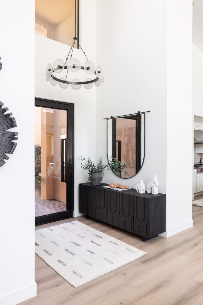 A modern entryway displays a black console, decorative vases, a round mirror, an abstract chandelier, and a white rug on light wood flooring. A glass door leads to the outdoor area—Living with Lolo.